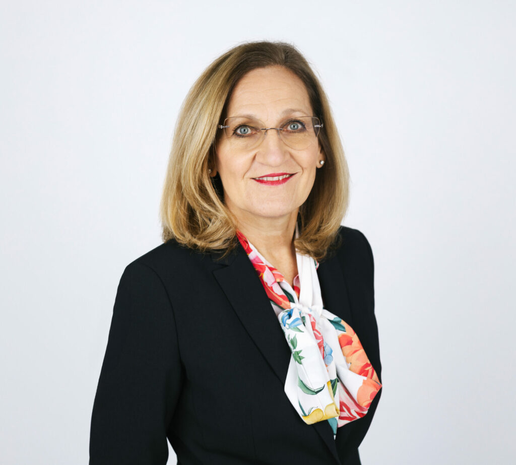 Frau mit schulterlangem blondem Haar, Brille, dunklem Blazer und gemustertem Halstuch vor hellem Hintergrund.
