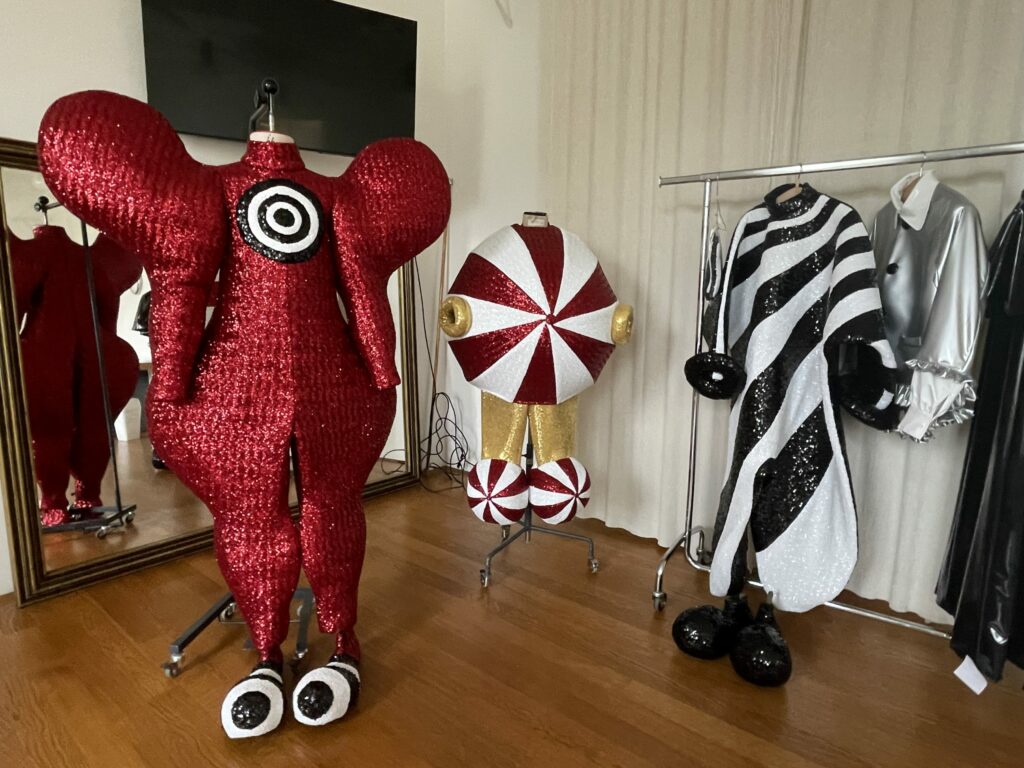 Drei bunte, auffällige Kostüme, eines rot mit großen Kreisen, eines rund gestreift und eines schwarz-weiß mit Glitzer, stehen auf Kleiderständern.